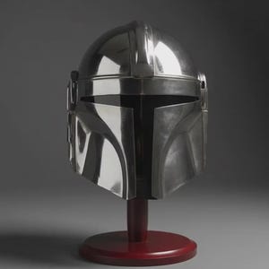 May include: A polished silver-coloured helmet with a smooth, reflective metallic finish, featuring a T-shaped visor opening and a central ridge along the top, mounted on a dark red circular wooden stand for display purposes.