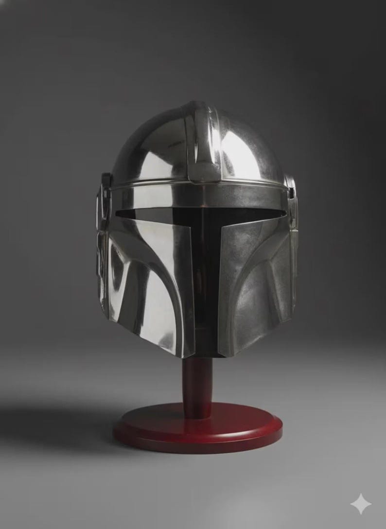 May include: A polished silver-coloured helmet with a smooth, reflective metallic finish, featuring a T-shaped visor opening and a central ridge along the top, mounted on a dark red circular wooden stand for display purposes.