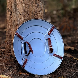 Op de afbeelding: Een zilverkleurig, cirkelvormig schild met concentrische ringen en rode leren riemen. Het schild heeft een centrale handgreep en extra riemen om het aan de arm te bevestigen. Het ontwerp doet denken aan een superheldenschild.