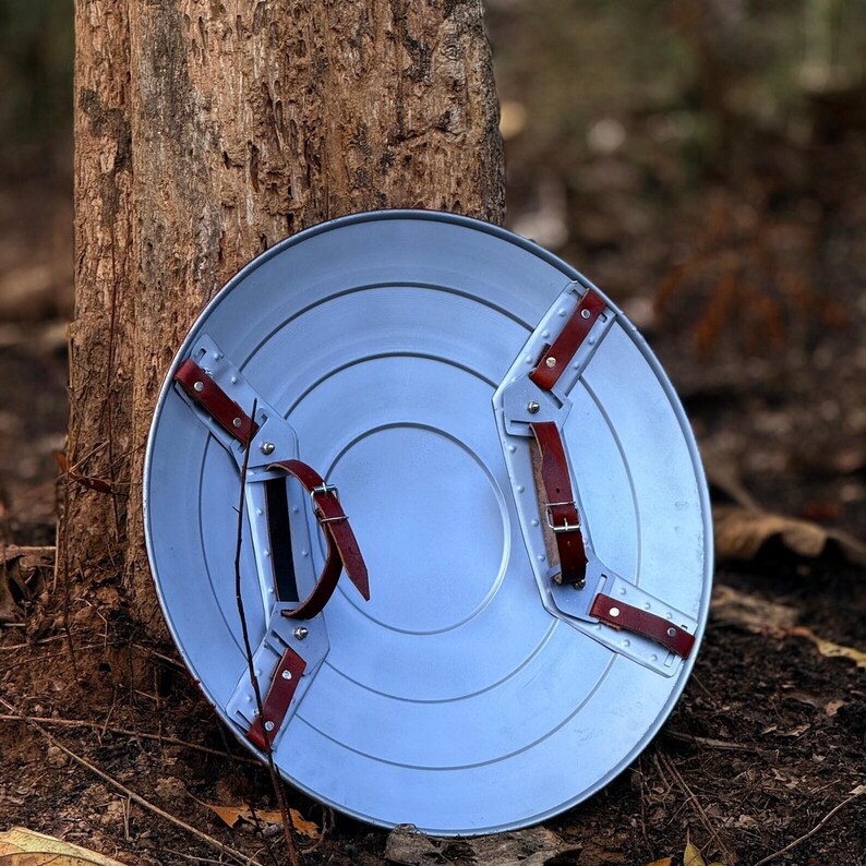 Op de afbeelding: Een zilverkleurig, cirkelvormig schild met concentrische ringen en rode leren riemen. Het schild heeft een centrale handgreep en extra riemen om het aan de arm te bevestigen. Het ontwerp doet denken aan een superheldenschild.