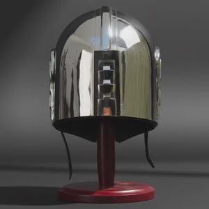 May include: A polished silver-coloured metal helmet with a smooth, reflective surface and a central vertical ridge. The helmet features a distinct rectangular cutout design on the front and includes thin black chin straps hanging down from the sides.