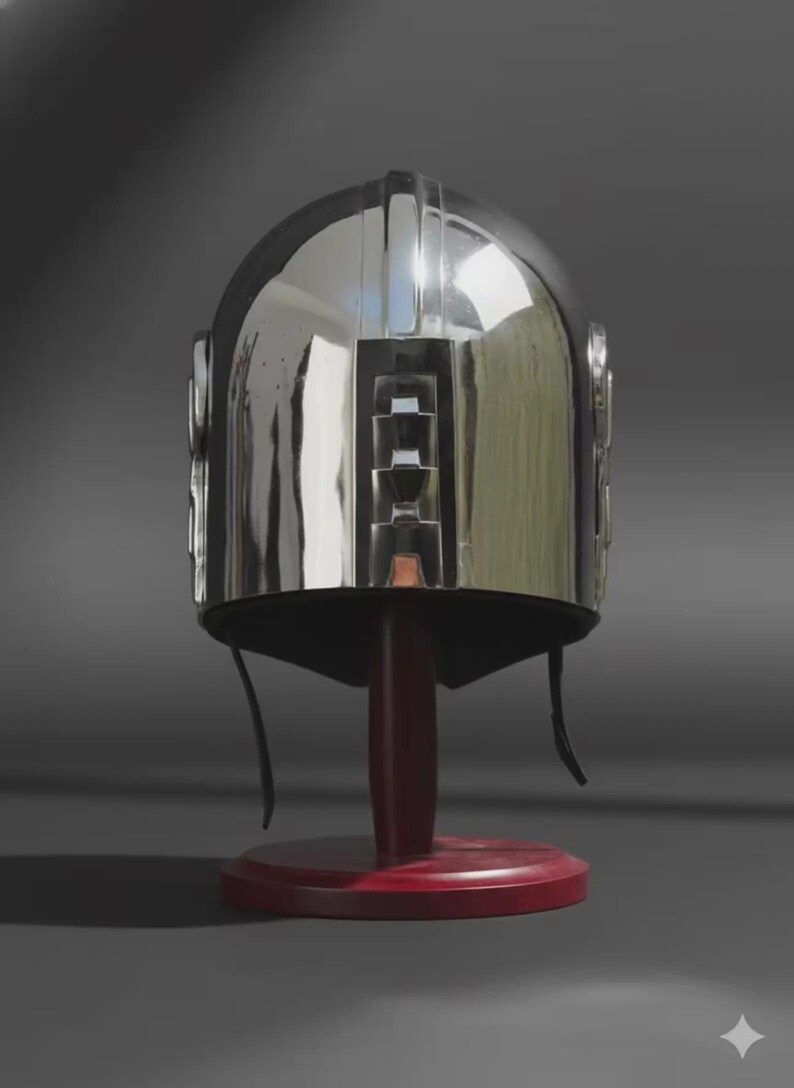 May include: A polished silver-coloured metal helmet with a smooth, reflective surface and a central vertical ridge. The helmet features a distinct rectangular cutout design on the front and includes thin black chin straps hanging down from the sides.