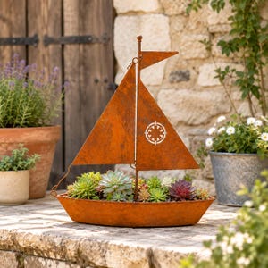 Puede incluir: Una maceta de metal color óxido con forma de velero, llena de varias suculentas verdes y moradas. El barco cuenta con un mástil con una vela triangular y una pequeña bandera en la parte superior. Se observa un diseño de brújula circular.