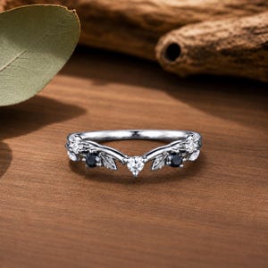 May include: A silver-toned V-shaped ring featuring a delicate leaf motif along the band. The ring is set with alternating round-cut clear white stones and round-cut black stones, creating a balanced contrast across the curved metal setting.