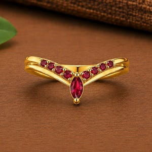 May include: A gold V-shaped ring featuring a central marquise-cut red gemstone flanked by six smaller round-cut red gemstones set along the band. The polished gold band follows a chevron silhouette, creating a symmetrical design with vibrant red stones.