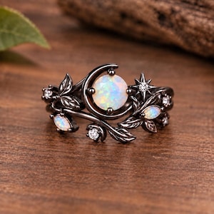 May include: A black metal ring featuring a central round iridescent opal stone set within a crescent moon shape, accented by smaller marquise-cut opals, round clear stones, and detailed leaf motifs along the band with a star-shaped setting.