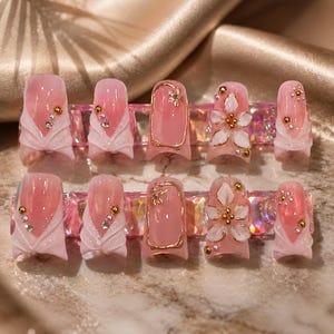 May include: A set of ten press-on nails featuring a translucent pink base with white sculpted accents, gold metallic outlines, and small floral embellishments. The design includes clear rhinestones and gold beads arranged on a pink gradient surface.