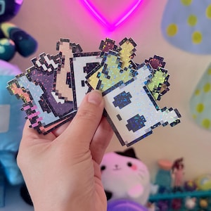 Op de afbeelding: Een set van drie regenboogkleurige holografische stickers met gepixelde spookfiguren. De stickers zijn in verschillende tinten paars, groen en geel.