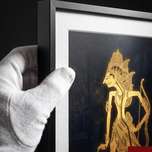 Puede incluir: Una pieza de arte de pared con marco negro, paspartú blanco y una impresión azul oscuro de una figura de marioneta de sombra Wayang Kulit dorada. La obra incluye el texto Harmony Soul en la esquina inferior derecha, con un estilo moderno.