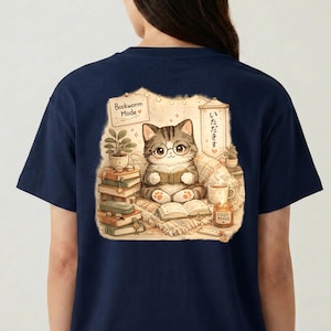 Könnte beinhalten: Ein marineblaues T-Shirt mit einem Grafikdruck einer getigerten Katze mit Brille, die ein Buch liest. Die Illustration zeigt Bücherstapel, eine Topfpflanze, eine dampfende Tasse, eine Kerze mit der Aufschrift Cozy Reads und den Text Bookworm Mode.