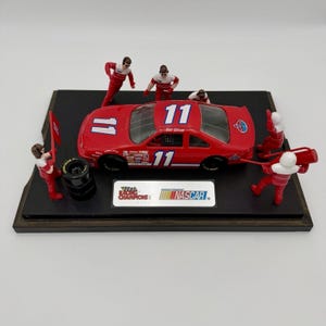 Könnte beinhalten: Ein rotes NASCAR-Modellauto mit der Nummer 11 und Bill Elliott-Schriftzug, präsentiert auf einem schwarzen Sockel mit vier Miniatur-Boxencrew-Figuren in rot-weißen Uniformen, einem Reifenstapel und einer Racing Champions NASCAR-Plakette.