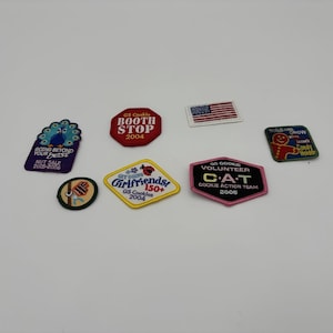 May include: A collection of Girl Scout patches with various designs and text. Patches include a peacock, an American flag, a red octagon with "Booth Stop," and a pink and black hexagon with "Cookie Action Team."