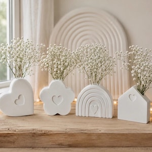 May include: A set of four white ceramic vases in various shapes including a heart, a flower, a rainbow, and a house. Each vase features a small embossed heart detail and is filled with delicate white baby's breath flowers, creating a cohesive display.