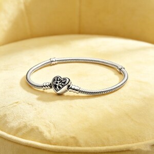 May include: A silver snake chain bracelet featuring a heart-shaped clasp. The clasp is decorated with an intricate tree design with small embedded stones. The bracelet has a smooth, flexible metallic texture and a polished silver finish.