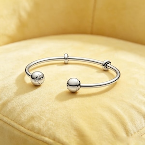 May include: A polished silver-tone open cuff bracelet featuring two large spherical end caps and two smaller sliding beads along the band. The spherical end caps are engraved with the text PANDORA, showcasing a sleek and metallic design.