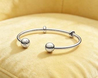 May include: A polished silver-tone open cuff bracelet featuring two large spherical end caps and two smaller sliding beads along the band. The spherical end caps are engraved with the text PANDORA, showcasing a sleek and metallic design.