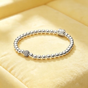 Könnte beinhalten: Ein silbernes Perlenarmband bestehend aus einer Reihe polierter Metallkugeln. Eine Perle ist mit kleinen klaren Steinen besetzt und eine zylindrische Perle ist mit dem Schriftzug PANDORA graviert, was ein einheitliches kreisförmiges Design ergibt.
