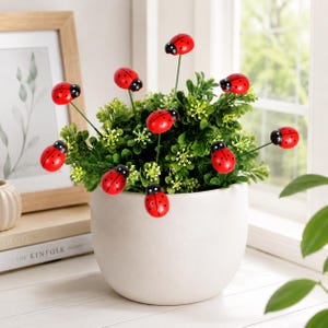 Peut inclure: Un pot en céramique blanche contenant une plante artificielle au feuillage vert luxuriant, décorée de plusieurs petites coccinelles rouges rondes fixées sur de fines tiges métalliques, avec des points noirs et une tête noire.