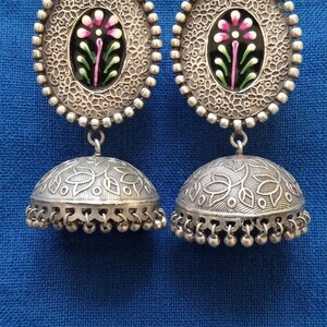 May include: A pair of silver-toned, ornate earrings. Each earring features a floral design in pink, green, and white on a black background, surrounded by a beaded border. The lower portion is a bell shape with intricate patterns and small dangling beads.
