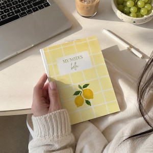 May include: A yellow notebook with a lemon illustration and the text "MY NOTES bella" is held in a person's hand. The notebook has a grid pattern. A laptop, pen, grapes, and a drink are in the background.