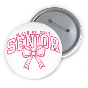Pode incluir: Um broche redondo com fundo branco e o texto rosa CLASS OF 2027 SENIOR arqueado sobre um desenho de linha rosa de um laço. A parte de trás do broche é de metal prateado com um mecanismo de alfinete horizontal para fixação.