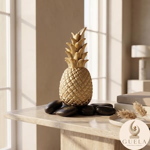 May include: A gold-colored pineapple sculpture with a textured, diamond-patterned body and pointed leaves at the top. The sculpture is surrounded by several smooth, dark gray river stones at its base. A circular logo with the text GUELA is visible.