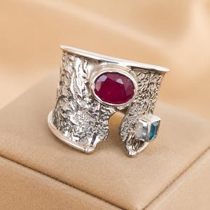 May include: A wide silver ring featuring a textured, uneven surface. The ring is set with a large, oval-shaped deep red gemstone and a smaller, square-cut blue gemstone positioned near the open end of the band.