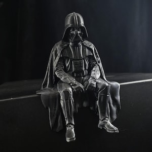 Op de afbeelding: Een matzwart beeldje van een zittend personage met een helm, een bedieningspaneel op de borst en een golvende cape. Het figuur zit in een rusthouding met de handen op de knieën en is voorzien van gedetailleerde bepantsering en textuur.