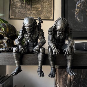 May include: Two detailed silver-toned Predator figurines in a seated position. One figure wears a smooth helmet, while the other displays a textured, organic head with dreadlocks. Both figures feature intricate armour plating and clawed feet.