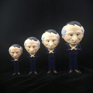 Puede incluir: Cuatro figuras de una persona mayor con cabeza grande, pelo gris y nariz prominente, que viste un traje azul oscuro y una pajarita amarilla. Cada figura está de pie con las manos juntas frente al pecho, dispuestas en orden de tamaño.