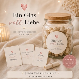 Op de afbeelding: Een doorzichtige glazen pot met een kurken deksel gevuld met kleine papieren rolletjes vastgebonden met touw. Het etiket vermeldt Ein Glas voll Liebe. Drie kleine kaartjes met Duitse tekst en hartjes staan naast de pot.