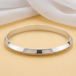 May include: A polished silver-tone bangle bracelet with a smooth, bevelled edge design. The circular metal band features a reflective, metallic finish that catches the light, creating a clean and simple aesthetic for this wrist accessory.