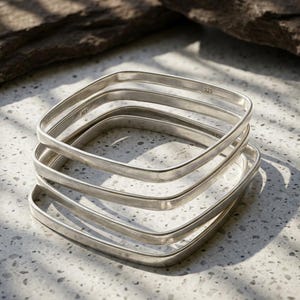 May include: A stack of four square-shaped silver bangles with a smooth, polished finish. Each bangle features a 925 hallmark stamp on the inner surface, indicating sterling silver composition. The bracelets are arranged in a slightly offset vertical pile.