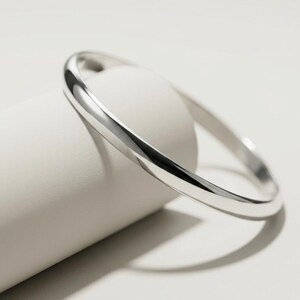May include: A polished silver-toned bangle bracelet with a smooth, rounded surface and a continuous circular shape. The metal reflects light, creating bright highlights along the curved edge of the band, showcasing a simple and clean design.