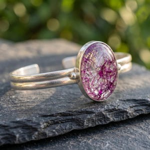 May include: A silver cuff bracelet featuring a prominent oval-shaped centerpiece. The centerpiece contains a clear, crackled glass-like material with vibrant magenta and purple inclusions embedded within the structure of the setting.