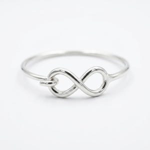 May include: A polished silver ring featuring a central infinity symbol design. The band is thin and circular, connecting to the continuous loop shape in the middle. The metal has a smooth, reflective finish, creating a clean and simple appearance.