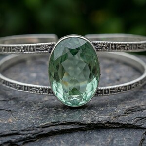 Op de afbeelding: Een zilveren armband met een grote, ovaal geslepen, lichtgroene edelsteen in het midden. De metalen band is verdeeld in twee parallelle armen, elk versierd met een ingewikkeld, in reliëf gemaakt bloemmotief over de gehele lengte.