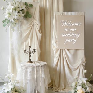 May include: A beige fabric backdrop display featuring a sign with the text Welcome to our wedding party. The setup includes a round table with a white lace cloth, a metal candelabra with three white candles, pearl strands, and white floral arrangements.