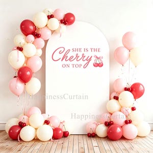 May include: A white arched backdrop featuring the pink text "SHE IS THE Cherry ON TOP" with a cherry illustration. The display is decorated with a balloon garland in shades of red, cream, and light pink, accented with small artificial cherries.