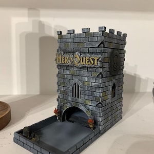 Pode incluir: Uma torre de dados HeroQuest cinza e preta, projetada para se assemelhar a um castelo. A torre apresenta o logotipo HeroQuest, um emblema de caveira e uma área de lançamento de dados. A torre tem uma textura semelhante a tijolos e inclui elementos decorativos.