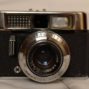 Pode incluir: Câmera Proniur 500 LK preta e prateada vintage com lente Voigtländer. A câmera apresenta um anel de lente prateado com configurações de abertura e um corpo de couro sintético preto. A lente é marcada 2.8/50.