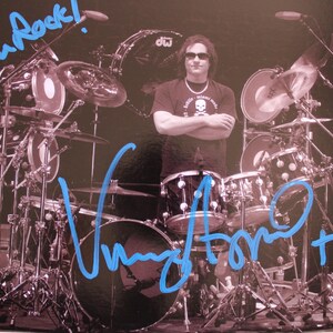 Puede incluir: Una fotografía en blanco y negro de un baterista sentado detrás de una gran batería con los brazos cruzados. La imagen presenta un autógrafo en tinta azul y el texto manuscrito "You Rock!" en la parte superior. Incluye una etiqueta de autenticidad.