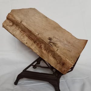 Puede incluir: Un libro antiguo con texto descolorido, apoyado en un soporte de metal oscuro. Las páginas del libro están envejecidas y manchadas, lo que sugiere su importancia histórica. El soporte tiene un diseño calado decorativo.