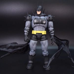 May include: A Batman action figure featuring a grey and black suit with spiked shoulder armour, a yellow utility belt, and a flowing black cape. The figure has a detailed muscular build, a cowl with pointed ears, and black gauntlets with grey stripes.