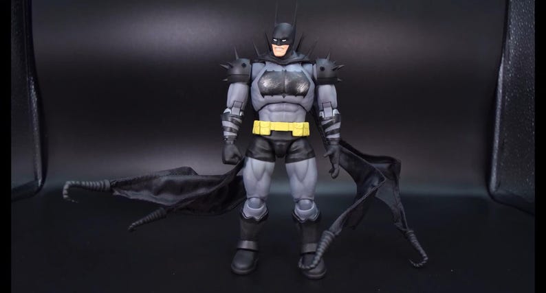 May include: A Batman action figure featuring a grey and black suit with spiked shoulder armour, a yellow utility belt, and a flowing black cape. The figure has a detailed muscular build, a cowl with pointed ears, and black gauntlets with grey stripes.