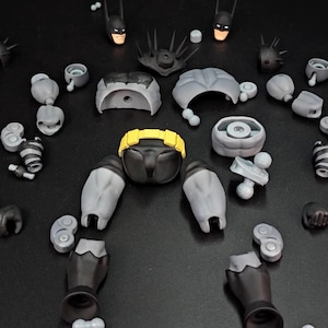 May include: A disassembled action figure kit featuring grey and black body parts, two interchangeable head sculpts with masks, a bright yellow utility belt, various hand attachments, a brown staff, and a metallic grey bat-shaped accessory.