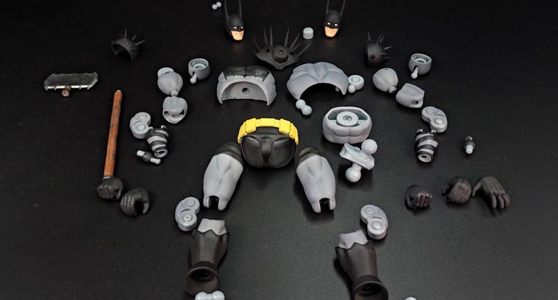May include: A disassembled action figure kit featuring grey and black body parts, two interchangeable head sculpts with masks, a bright yellow utility belt, various hand attachments, a brown staff, and a metallic grey bat-shaped accessory.