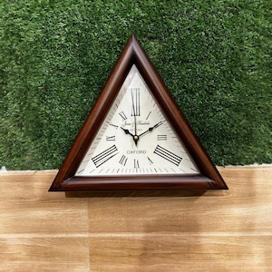 Puede incluir: Un reloj de pared triangular con marco de madera marrón oscuro y esfera de color crema. El reloj presenta números romanos negros, manecillas negras para las horas y los minutos, y el texto Jones & Hendricks EST 1925 y OXFORD en la esfera.