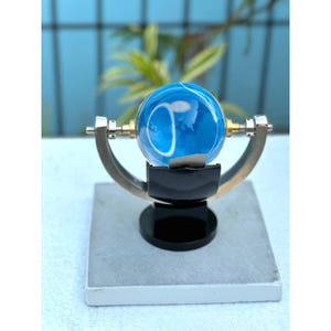 May include: A blue glass sphere with white swirling patterns mounted on a metallic gimbal frame. The sphere is held by a black support structure attached to a square base. The metallic frame features brass-colored accents at the pivot points.