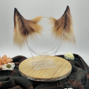 Puede incluir: Un par de orejas de animal de piel sintética puntiagudas con un degradado de marrón oscuro en las puntas que transiciona a tonos beige claro y crema en la base. Las orejas están unidas a una estructura de diadema de alambre fino.